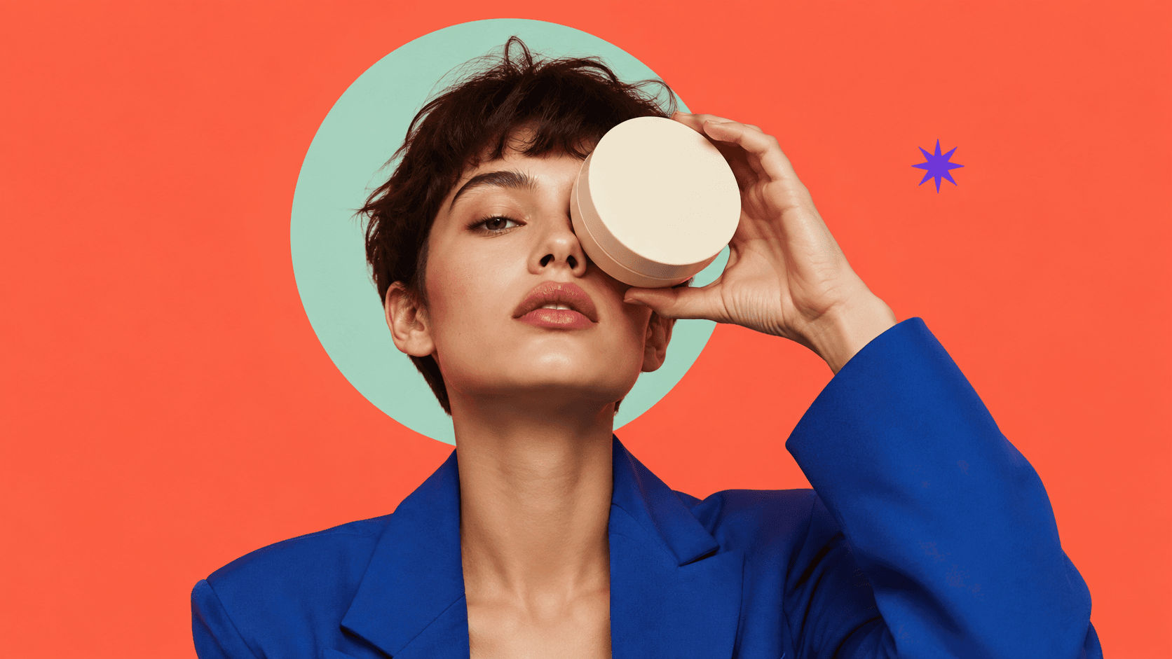 A model in a blue blazer on coral, holding a cream product jar in front of one eye